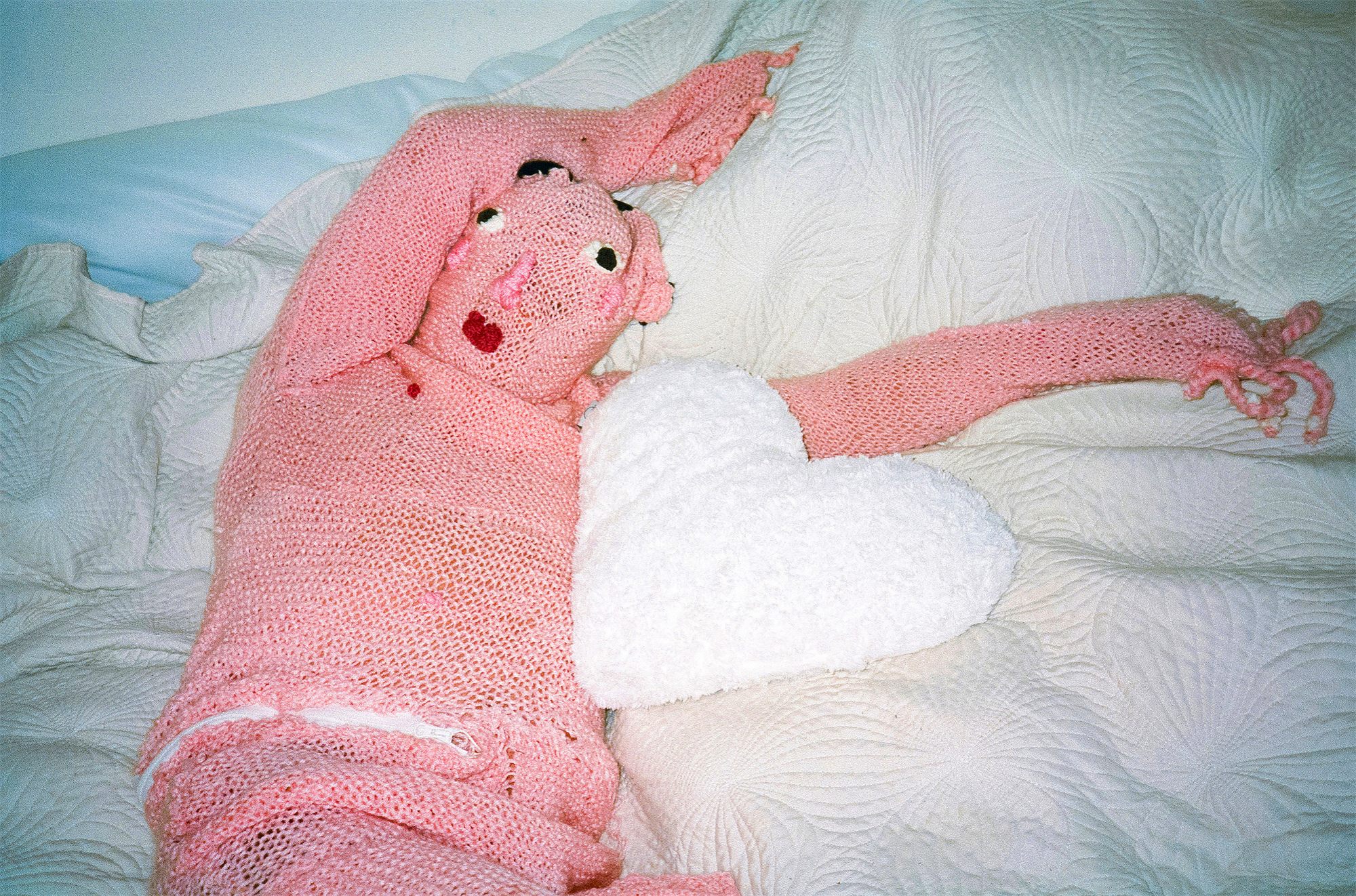 Éva Ostrowska, Divine, pink knitted figure on a white bed embracing a white heart cushion, photographic performance