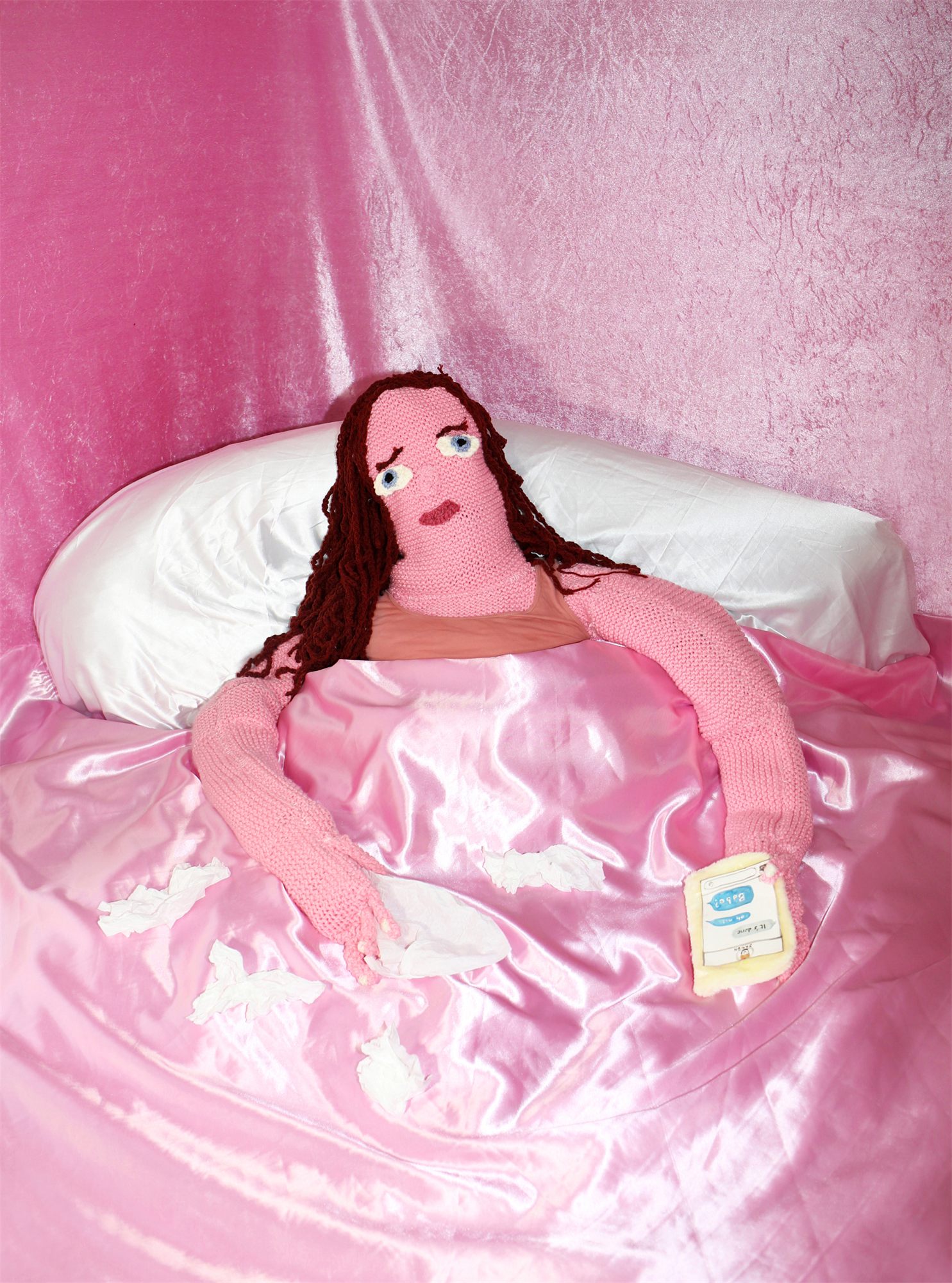 Éva Ostrowska, Ghosted, crying knitted figure with a knitted phone in a pink satin bed, photographic performance