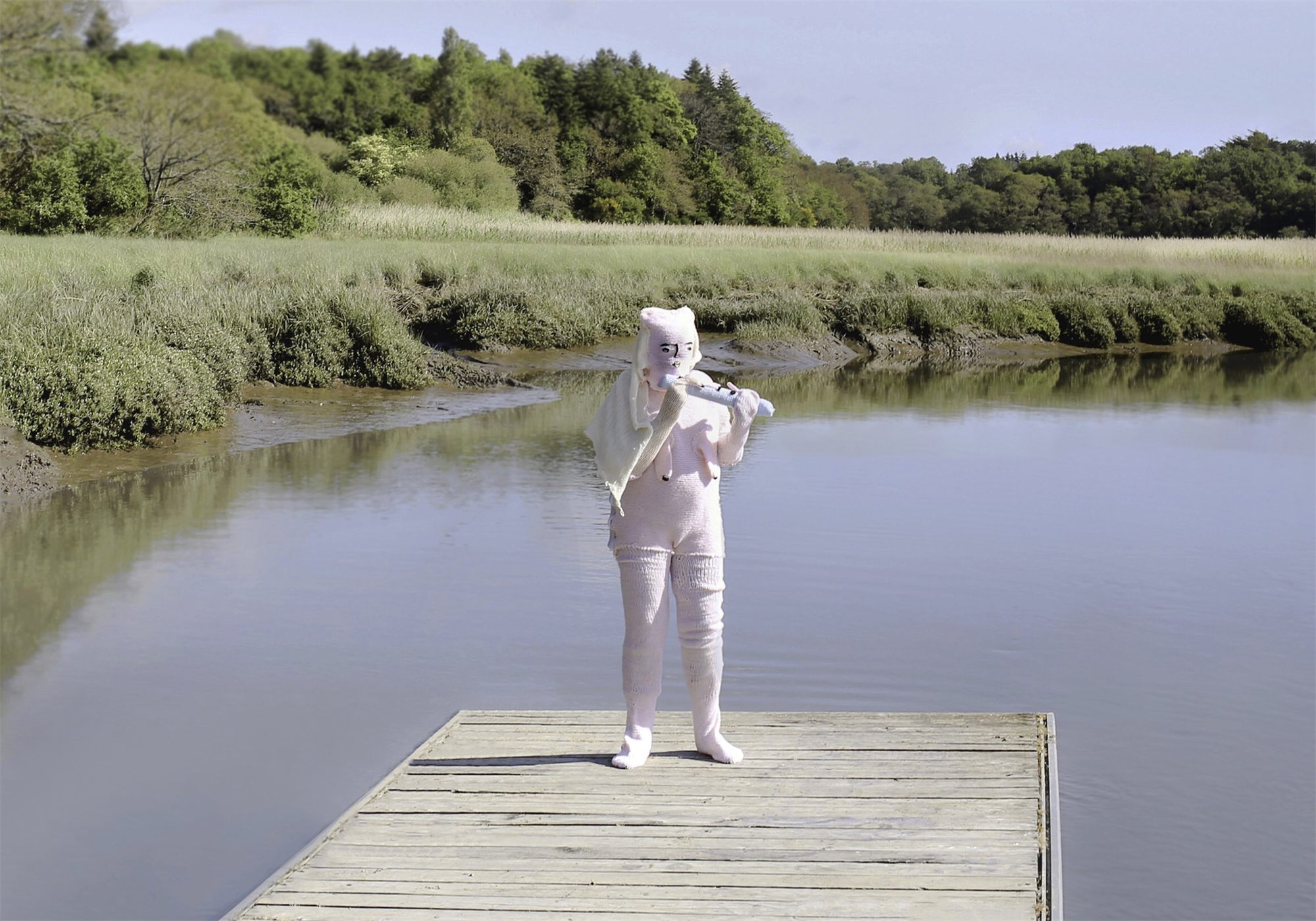 Éva Ostrowska, Pan, pale pink knitted figure playing a knitted flute on a wooden pontoon over still water, photographic performance