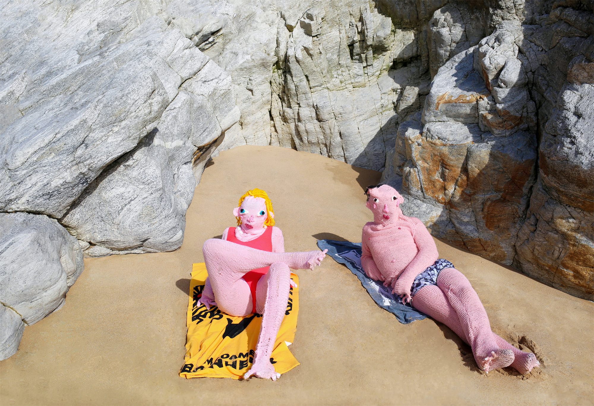 Éva Ostrowska, You and I, two pink knitted figures sunbathing on towels between pale rocks, photographic performance
